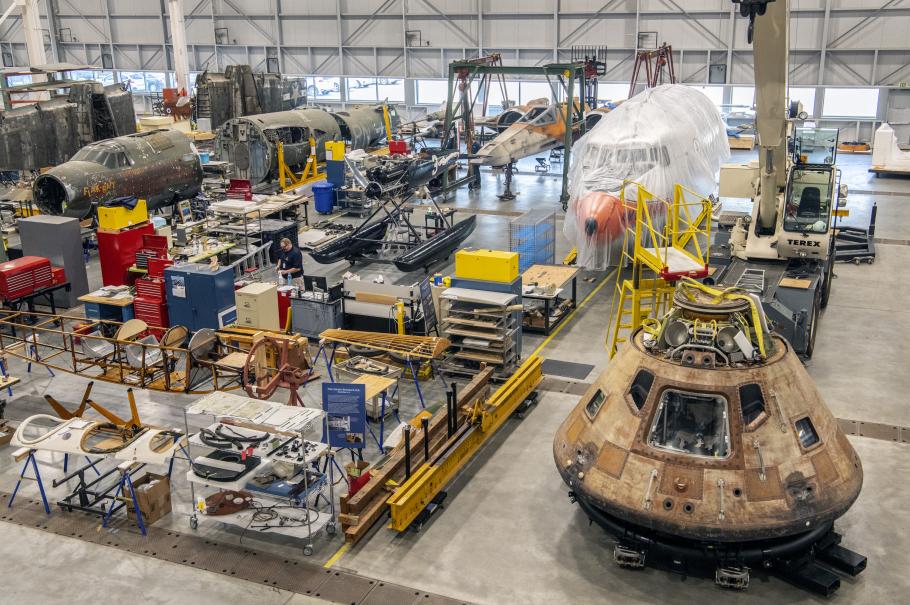 A hanger filled with air and spacecraft. In the foreground is the Apollo 11 Command Module, in the background is a WW2 plane labeled Flak-Bait. 