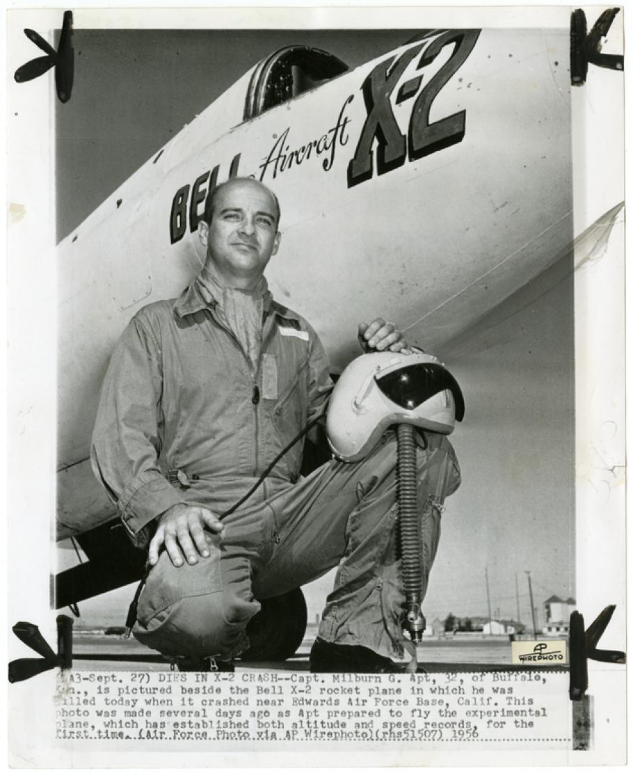 A man kneels in a flight suit in front of the nose of an aircraft which has Bell Aircraft X-2 written on the side. He balances a helmet on his knee.