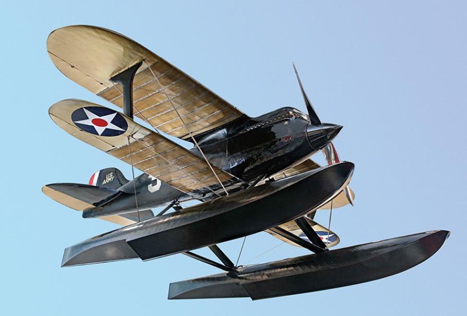 Low angle view of the James H. Doolittle's Curtiss R3C-2 racer, a black plane with a beige wing above and below the body. Below the second wing are long black floats for landing.