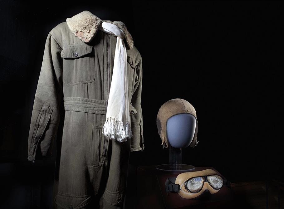 A olive flight suit ( a jumpsuit-like garment) with a fur collar and a white scarf, as well as tight fitting leather helmet and goggles stand against a black background.