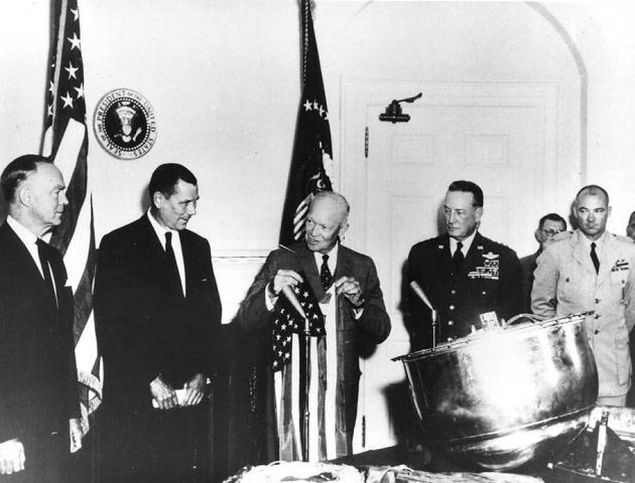 A group of 5 men stand around a table with a large metallic drum shaped object on it.