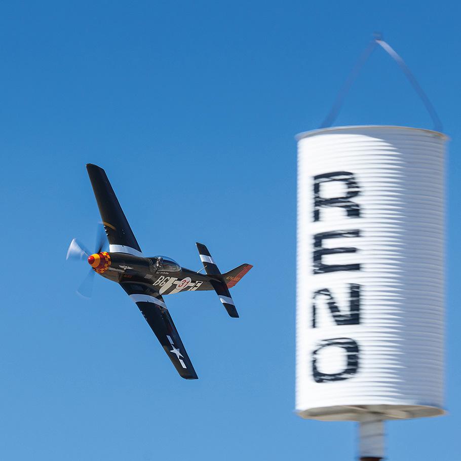 Speedball Alice, a restored dark green P-51D airplane with white stripes, makes a tight turn around a pylon with a white barrel shaped sign reading "Reno" on the top of it. Its propellor is spinning so fast its a whir, reflecting the aircraft's speed.