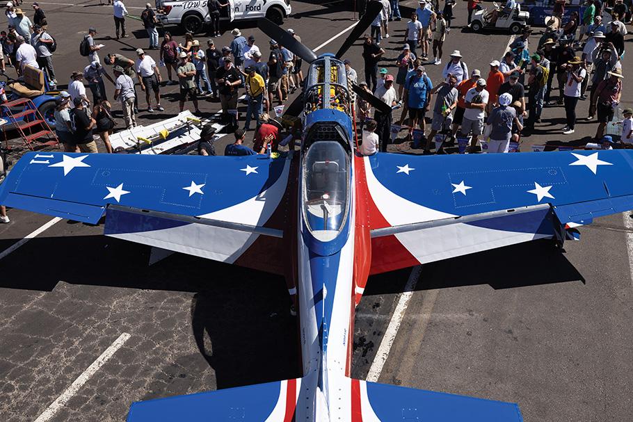 The patriotically painted red, white, and blue airplane, Miss America, is surrounded by a crowd of race fans as it sits on the tarmac on a bright sunny day in Reno.