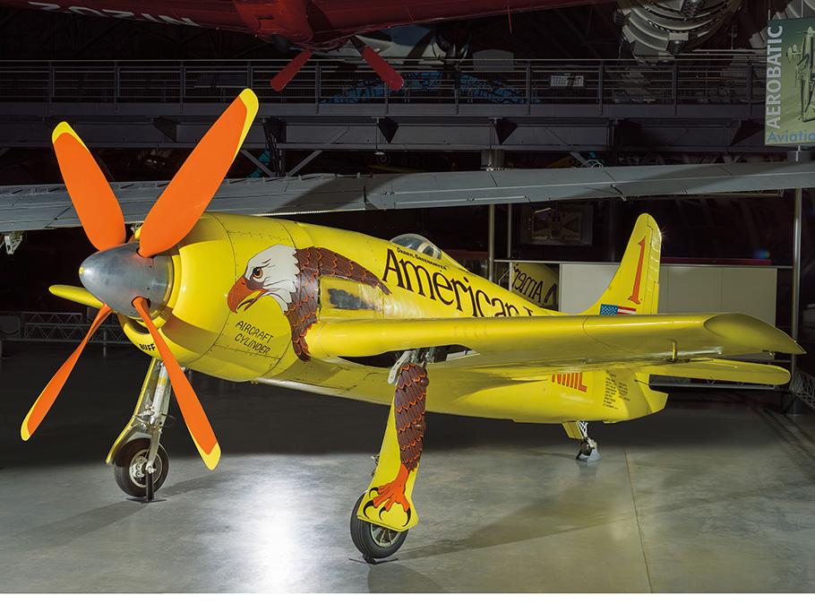 A yellow Grumman F8F-2 Bearcat airplane sits in the Boeing Aviation Hangar at the National Air and Space Musuem's Steven F. Udvar-Hazy Center. The aircraft, named Conquest I, has a painting of an American Eagle covering it's side, with the bird's leg and claw extending downward on the right front wheel. The aircraft has a large orange propellor on its nose. 