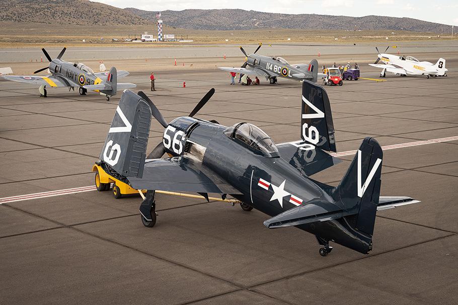 A Korean-War era Grumman F8F Bearcat, a grey airplane with a propellor on its nose, sits parked among other aircraft, with its wing tips positioned upwards.