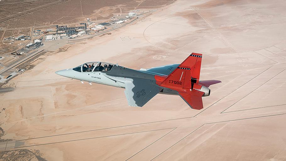 The first T-7A Red Hawk airplane soars over Edwards Air Force Base. True to its name, the most distinctive feature is its bright red tail fins that stand out from the metallic gray color of the rest of the aircraft.