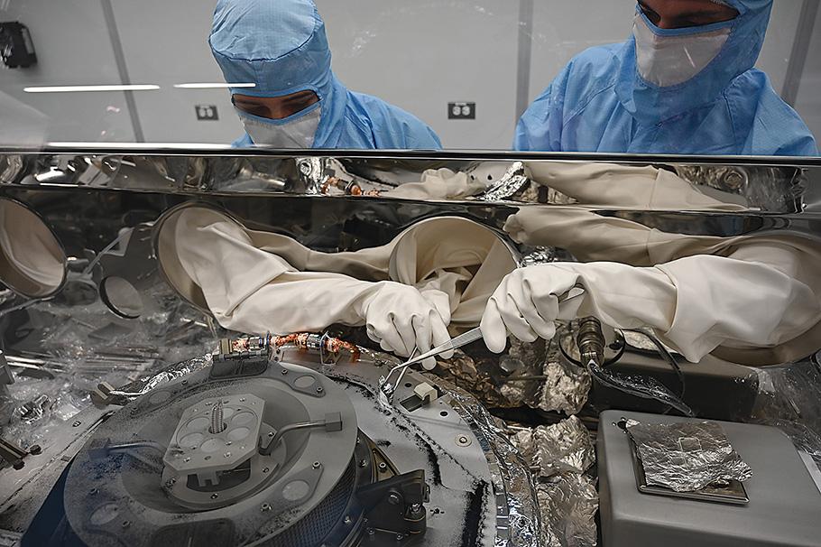 Peering through the glass casing of a glovebox, two masked technicians wearing light blue clean room suits with white gloves use small metal tools to extract asteroid samples from the OSIRIS-REx capsule.