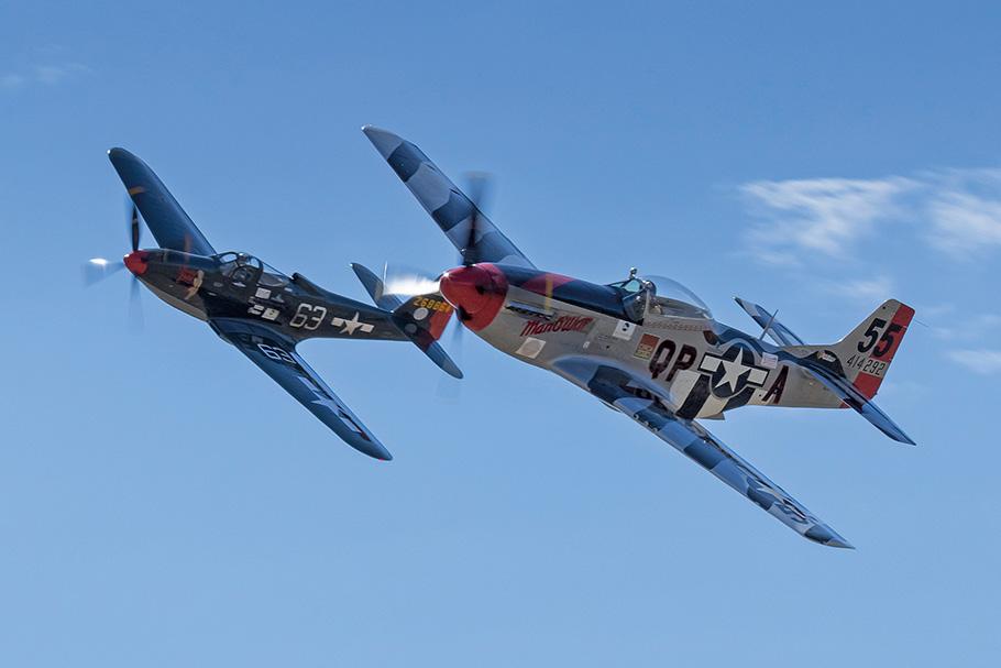 The dark blue P-51D Man O War plane races through the sky alongside Pretty Polly, a P-63C plane with a grey fuselage and blue-striped wings.