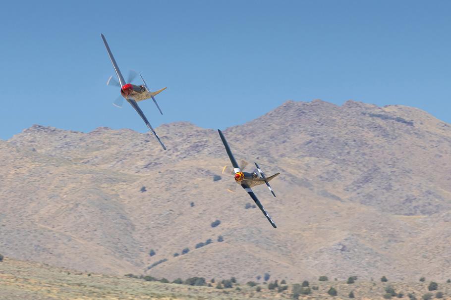 Two P-51D airplanes are tilted to the right as they make a run to the grandstands, the mountains of the Reno desert in the background.