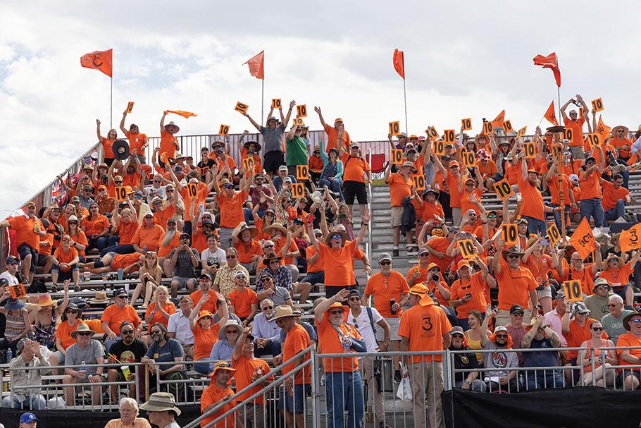 In a grandstand, a group of fans wearing orange t-shirts and caps are cheering as they watch the air races. Many hold up orange scorecards with the number 10. 
