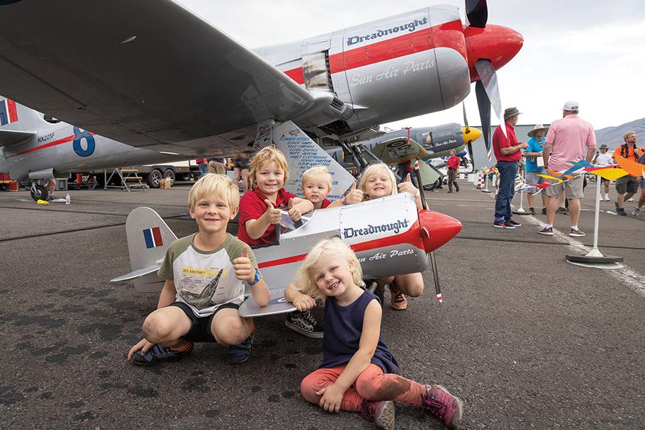 A group of blond-haired toddlers play with a kid-size version of Dreadnought, a modified Hawker Sea Fury airplane with a distinctive red strip along its body. The full-sized plane is seen parked in the background.