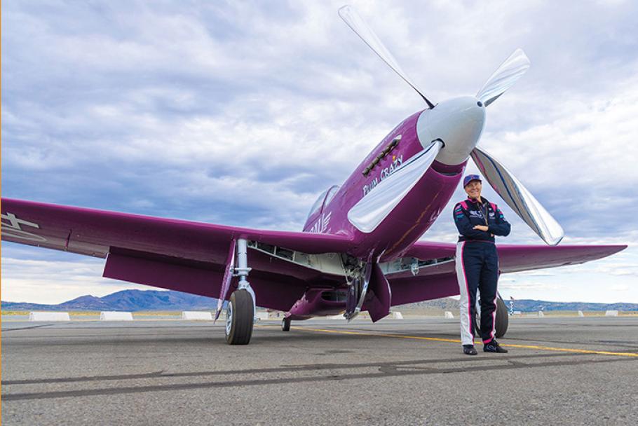 A woman wearing a blue-and-white flight suit (pilot Vicky Benzing) poses in front of Plum Crazy, a dazling restored P-51 airplane painted in distinctive purple with chrome propellors.