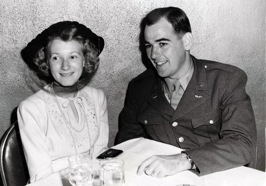 A white woman and a white man--both in their early 20s and both smiling--sit at a table. The man, a pilot, is wearing a military uniform with a set of wings pinned next to his lapel. The woman is wearing a white ensemble and a double strand of pearls.