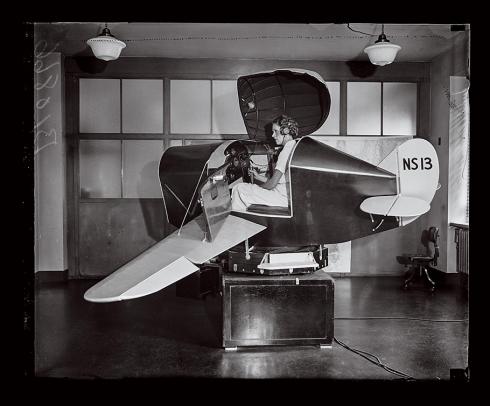 A woman sits indoors in a small, stubby aircraft that could be mistaken for a coin-operated child’s ride. This is a Link Flight Simulator, which trained more than 500,000 U.S. pilots.