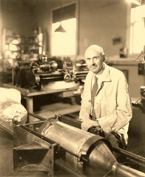 Informal pose of a bald mustachioed man, Dr. Robert H. Goddard, sitting in front of a rocket under construction in his workshop.