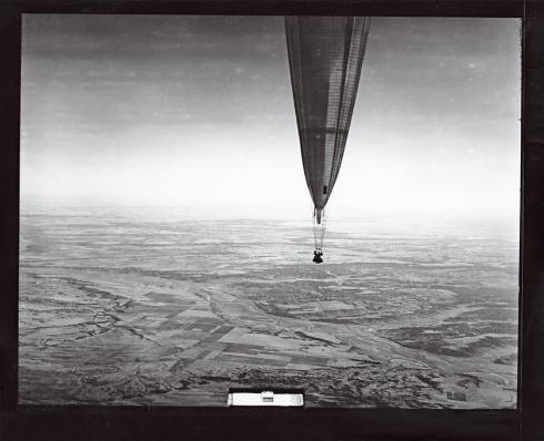 A black and white photograph of a balloon with a gondola floating high above a landscape.