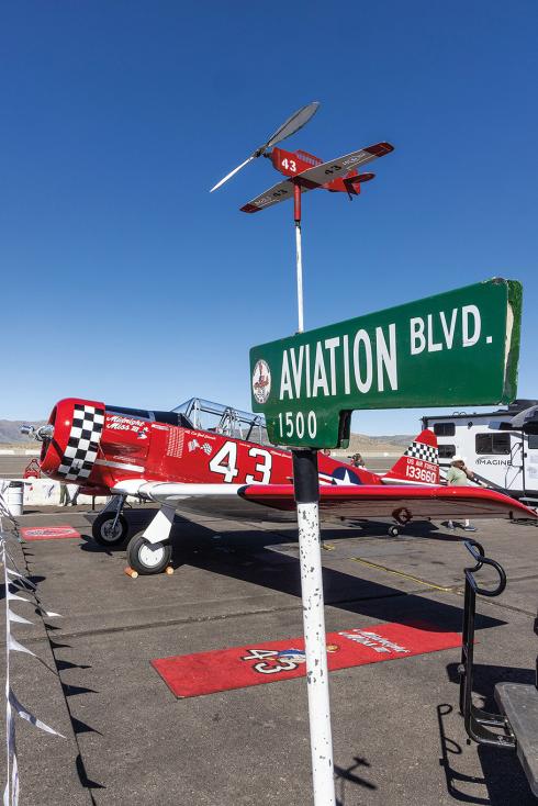 A red and black-checkered T-6 Texan airplane is parked near a street with a green sign saying, "Aviation Blvd." Above the street sign is a small windmill shaped like the T-6 Texan with a matching paint job. 