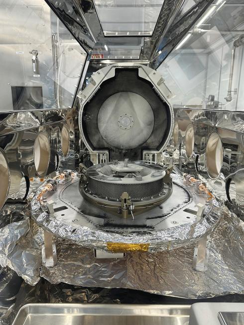 The OSIRIS-REx capsule sits inside of a scientific room with white sterile walls and metallic instruments. The capsule itself is circular, and opens at the mid-section like a waffle iron. 