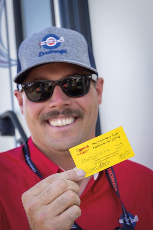 Steven Koewler, a middle-aged man wearing a red shirt, grey cape, and sunglasses, smiles as he holds up a bright yellow card.