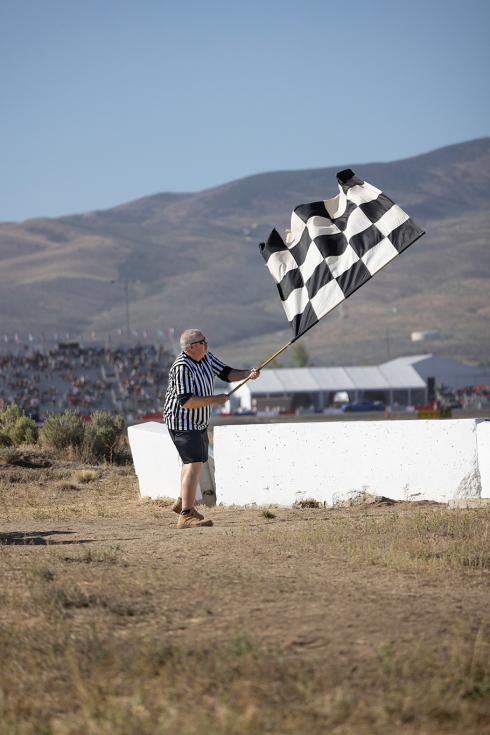 A senior man weaing a referee-style pinstripe shirt waves a large black-checkered flag on a pole on a sunny desert day at the races.