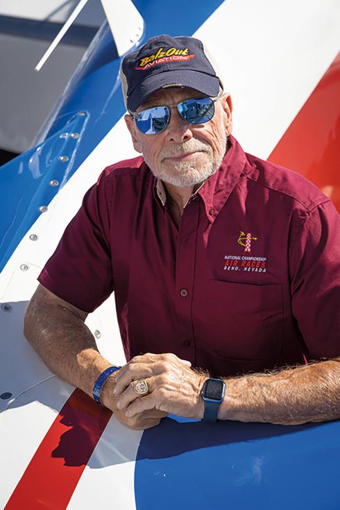 Alan Preston is a senior man wearing a red shirt, a dark grey cap, and mirrored sunglasses stares at the camera.