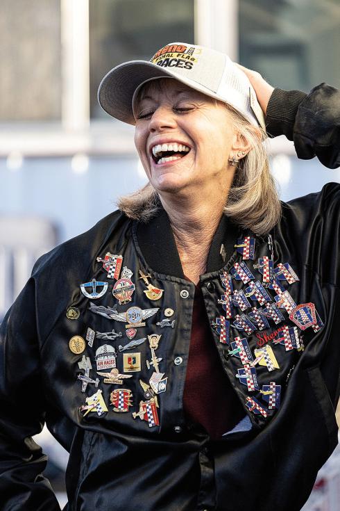 A middle-aged woman with shoulder-length blonde hair laughs while wearing a black jacket covered almost completely in souvenir air racing pins.