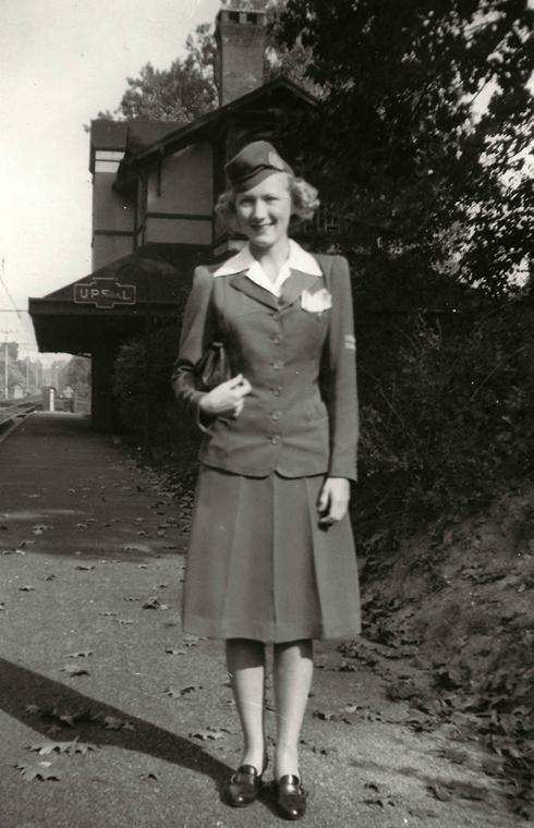 A smiling white woman in her early 20s stands in front of a train station with a sign that reads: Upsal. She is wearing a 1940s-era stewardess uniform: white blouse, buttoned jacket, pleated skirt, and matching hat.