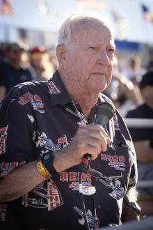 Sandy Sanders is a senior man wearing an air-race themed Hawaiian shirt. He holds a microphone while speaking. 
