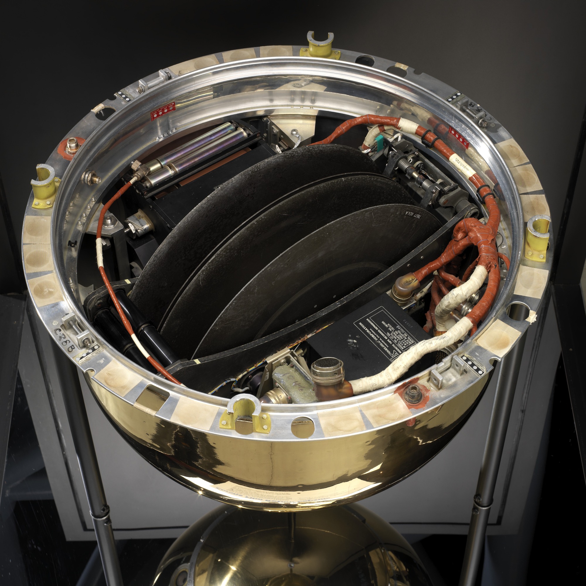 Bucket-shaped, gold-colored capsule and electronics, two black metal takeup spools; with metal cover and modified ring housed in black wooden case with black felt and clear plexiglas.