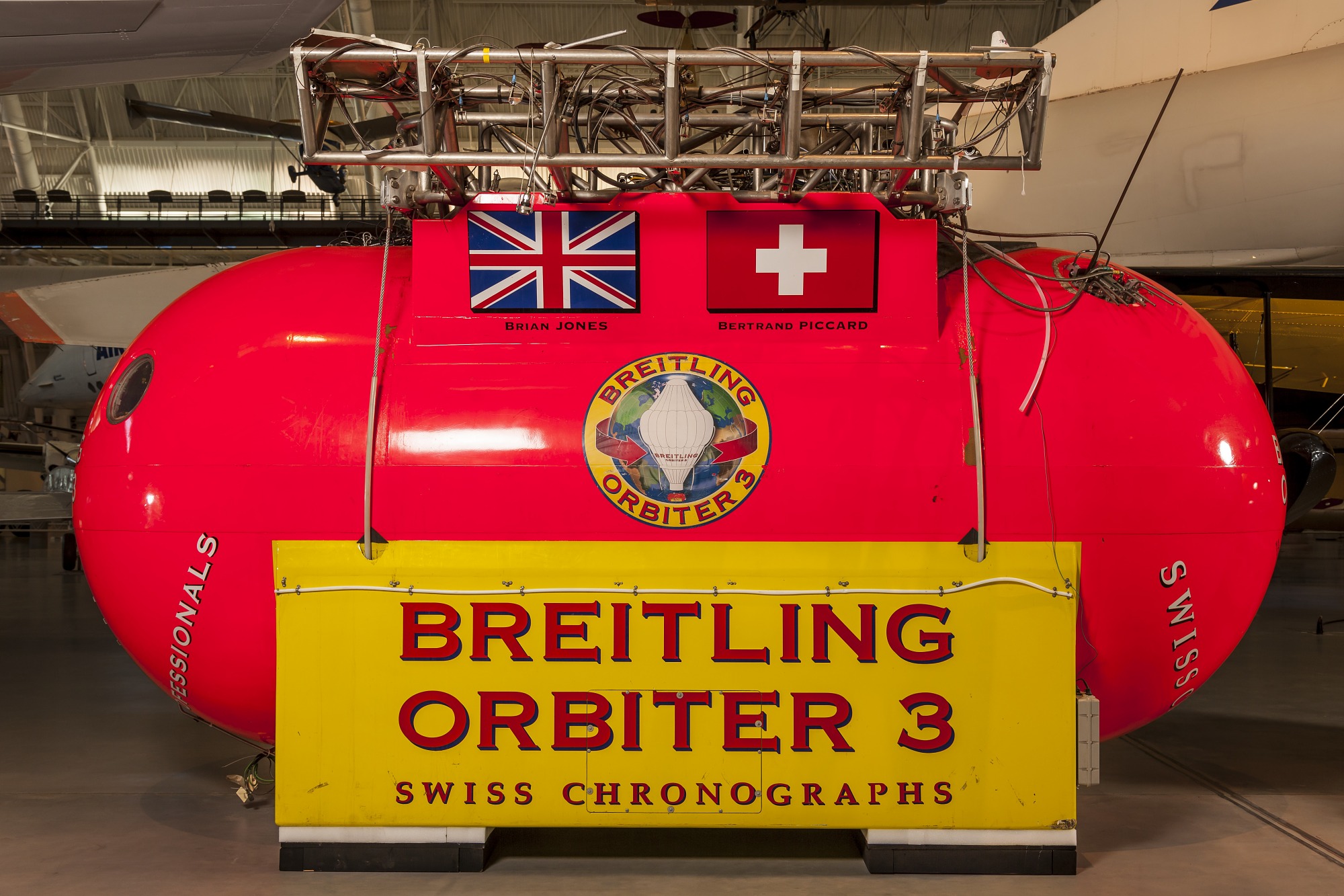 Bright red and yellow metal structure that is the gondola used to carry people and equipment during a balloon ride. It has the inscription, Breitling Orbiter 3. Attached to the metal structure are silver tanks. 