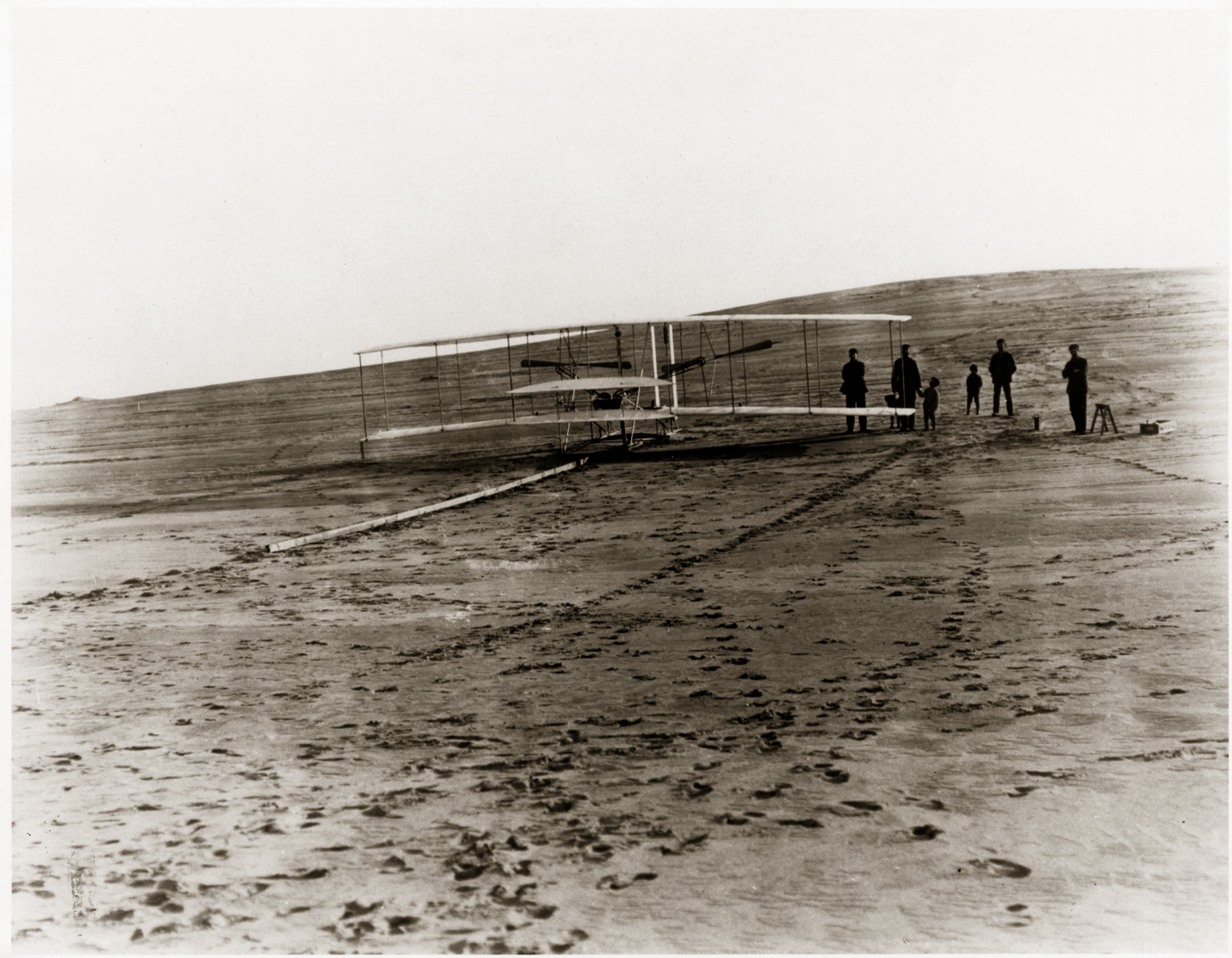 A biplane sits on a long wooden rail on the ground. A group of people stand behind it.