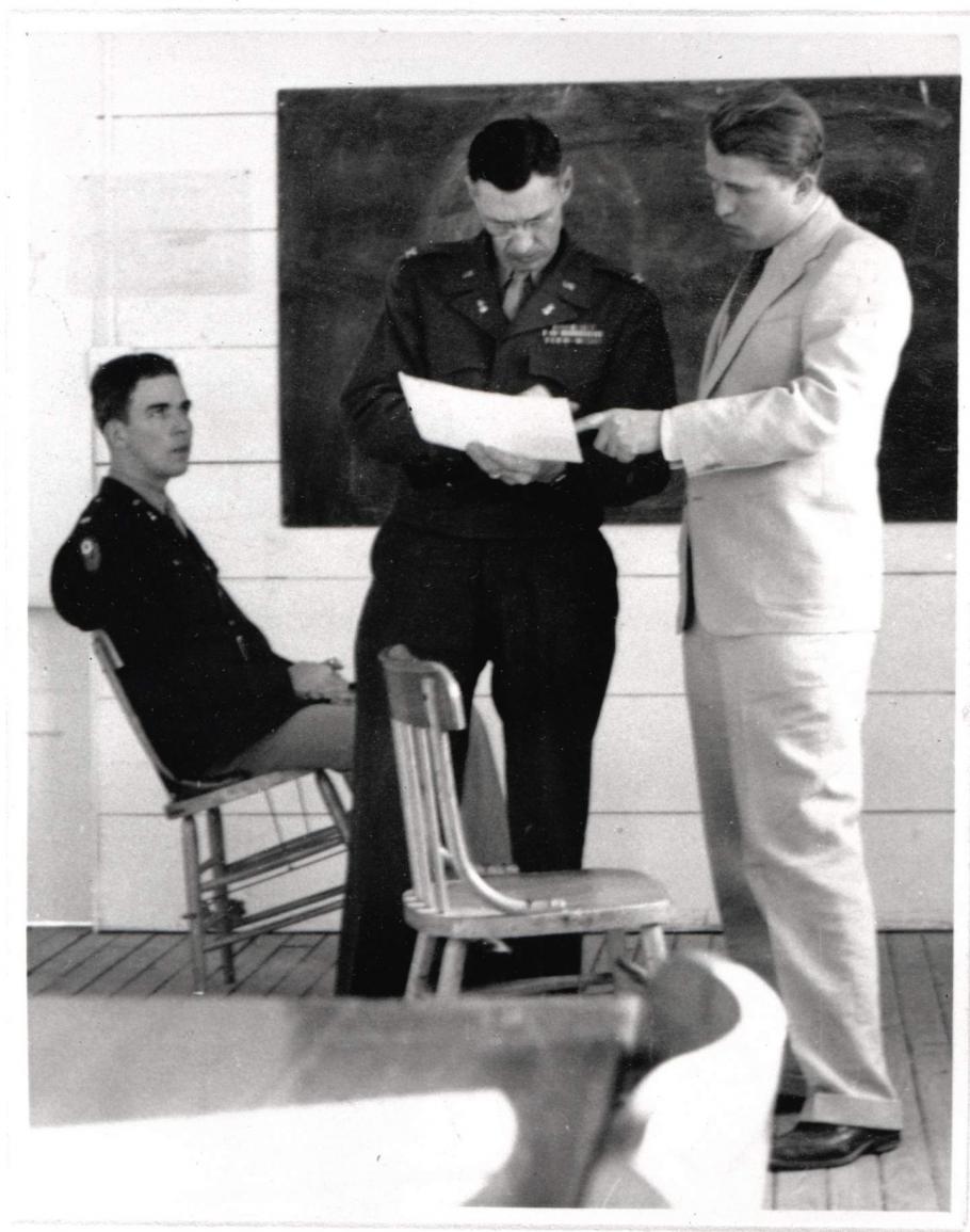 One man sits in a chair while two men stand up and look at a paper. 