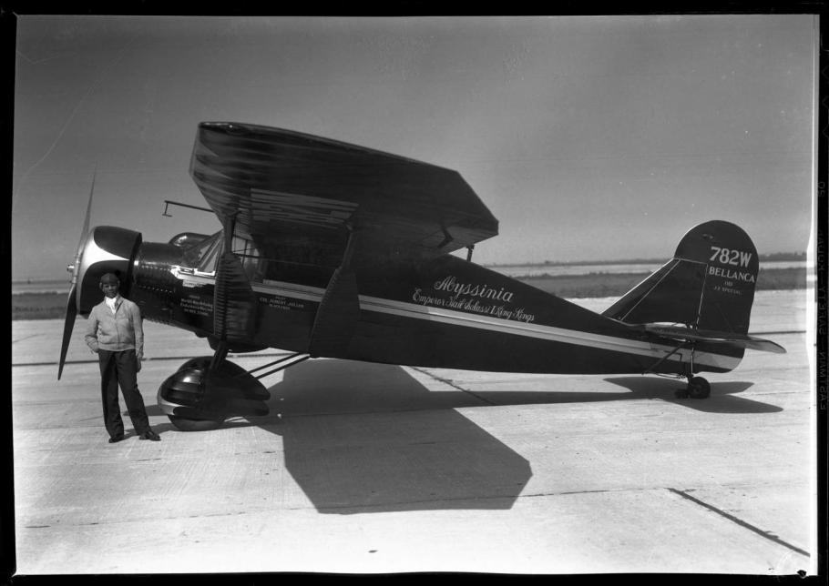 Black and white left side view of monoplane painted black with white stripe down middle. Cursive handwritten text in center of plane reads: "Abyssinia Emperor Haile Salassi I King of Kings." Tail text "782W/Bellanca" Black man by nose right hand in pocket