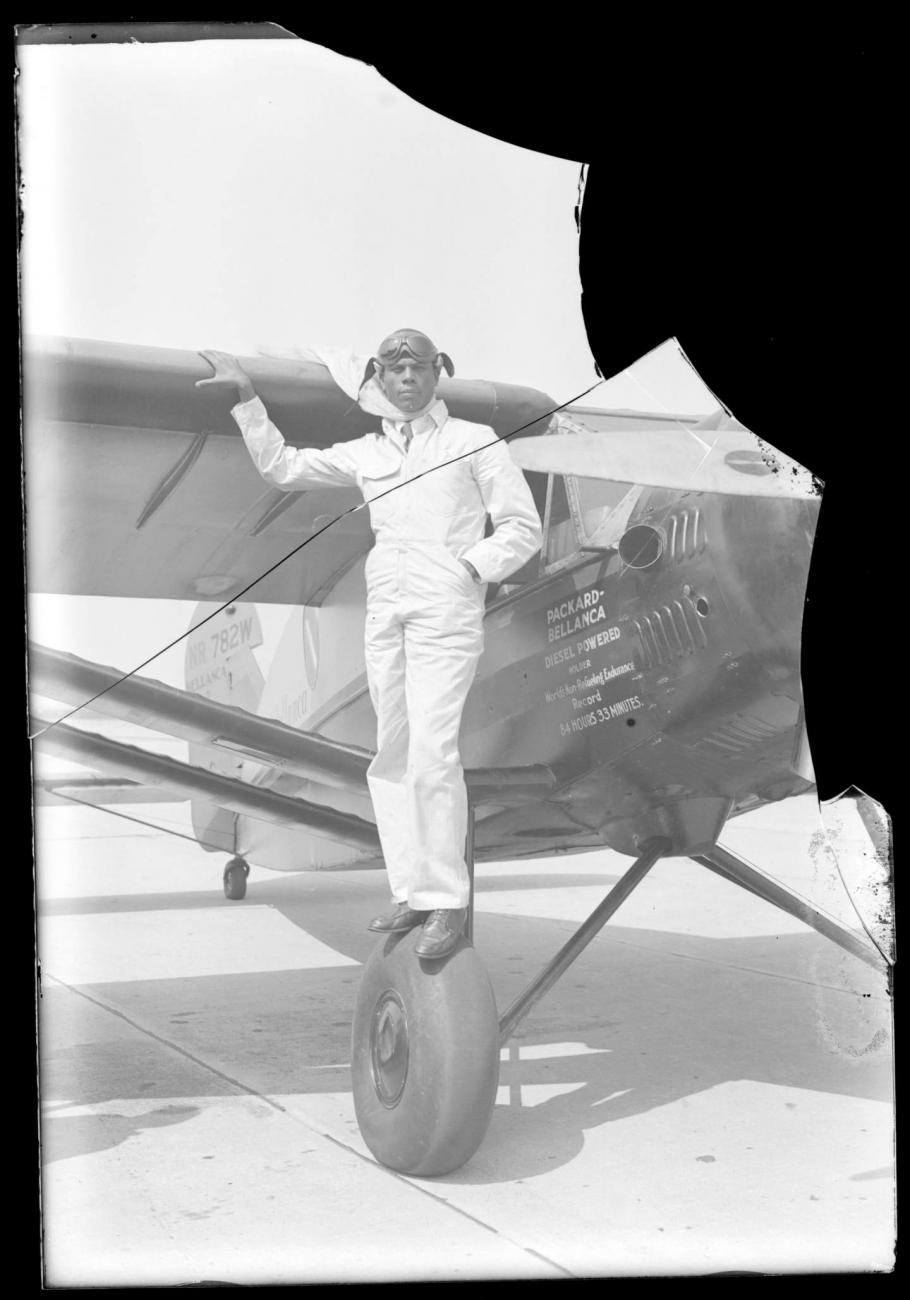 Three quarter right view of monoplane. Black man in white flight suit, poses standing on the right front wheel, holding wing in right hand.  Text under window reads: "Holder World’s Non-Refueling Endurance Record 84 Hrs 33 Mins" cracked upper right corner