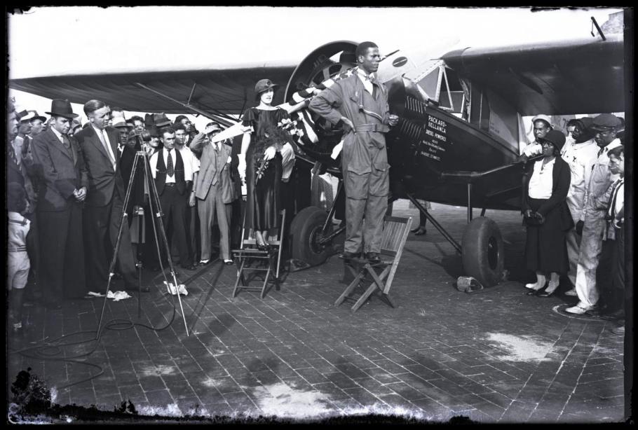 Black and white photo of Black man in flight suit standing on folding chair in front of left side of monoplane. Woman stands behind him holding champagne bottle. Crowd in the background with camera on tripod to the left.