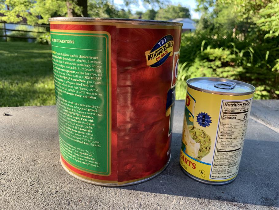 Two cans sit on table. (left) Large 3 quart can of tomatoes with red label (right) Smaller 15.5 ounce can of artichoke hearts with yellow label