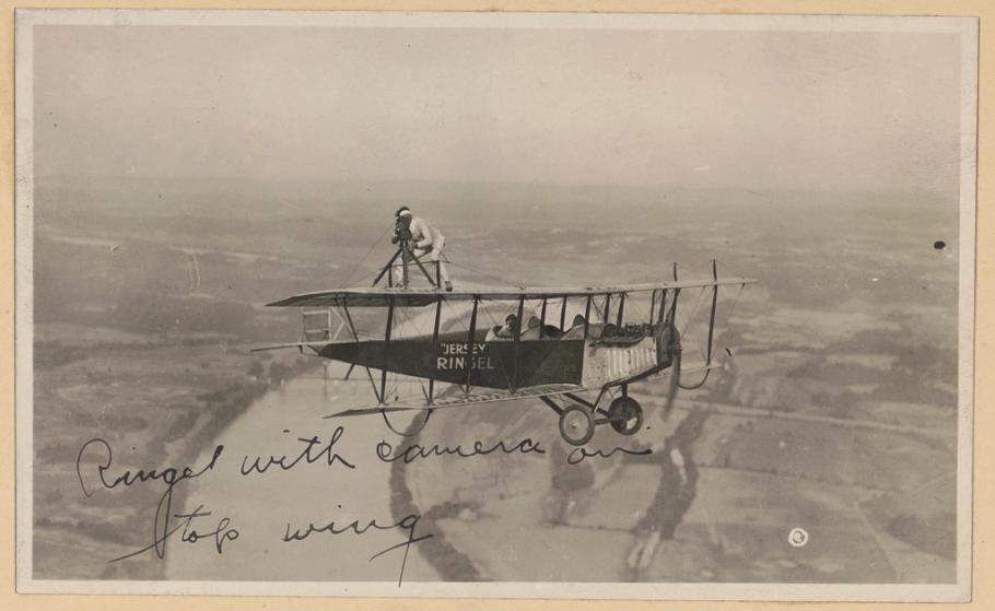 A man performing a barnstorming act standing on the wing of an airplane holding a camera