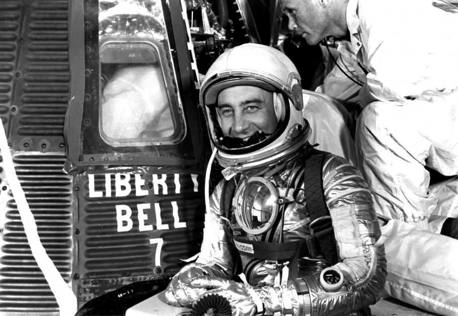 man in silver spacesuit smiles in front of space capsule that says LIBERTY BELL 7