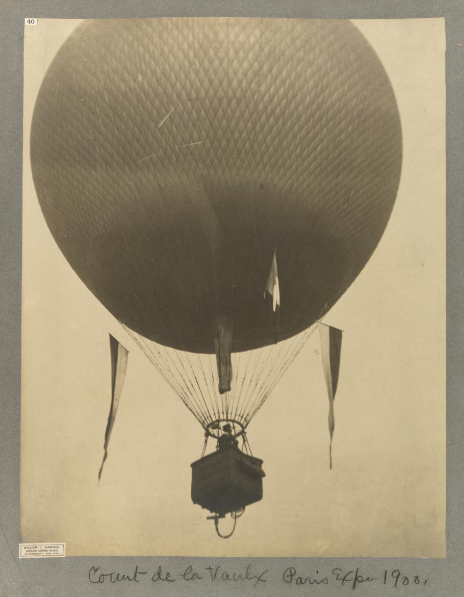 Black and white photograph of spherical hot air balloon, with man in basket, three flags hang off the ropes