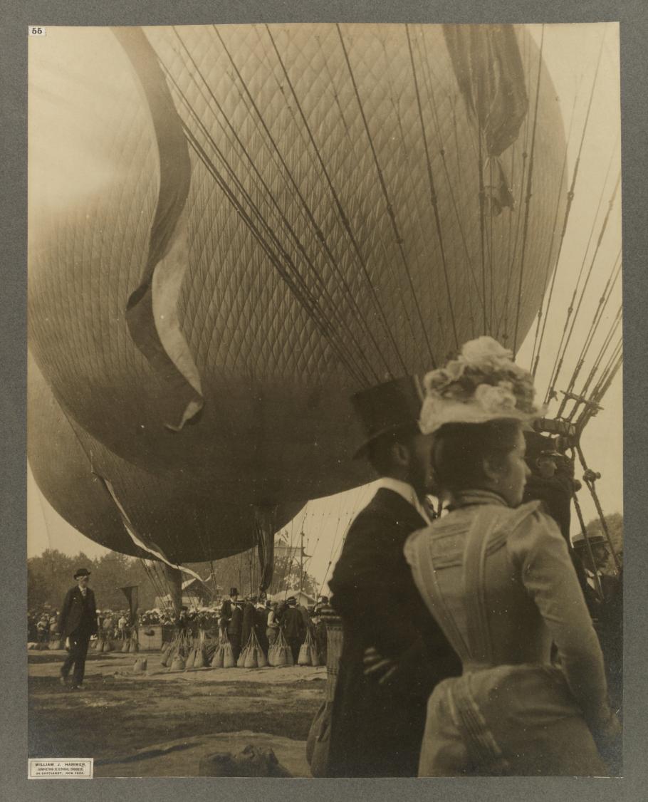 Black and white photo of the backs of a man in a suit and top hat and a woman in a white dress with floral hat with hot air balloons in the background