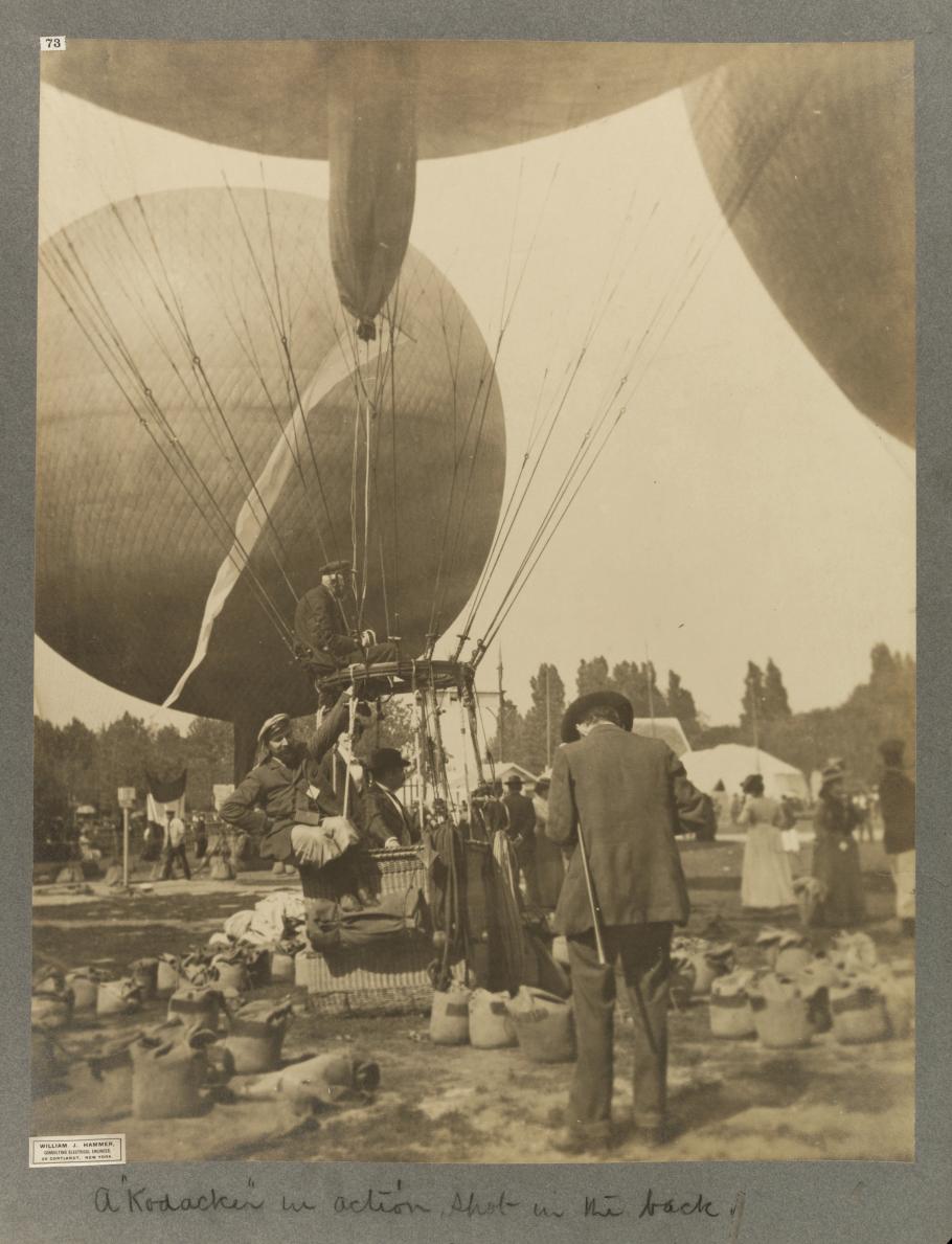 Black and white photograph of a man's back. We can presume that he is using a camera to take a photograph of the three men in the basket of a hot air balloon--the man on the left is partially hanging out of the basket on a rope.