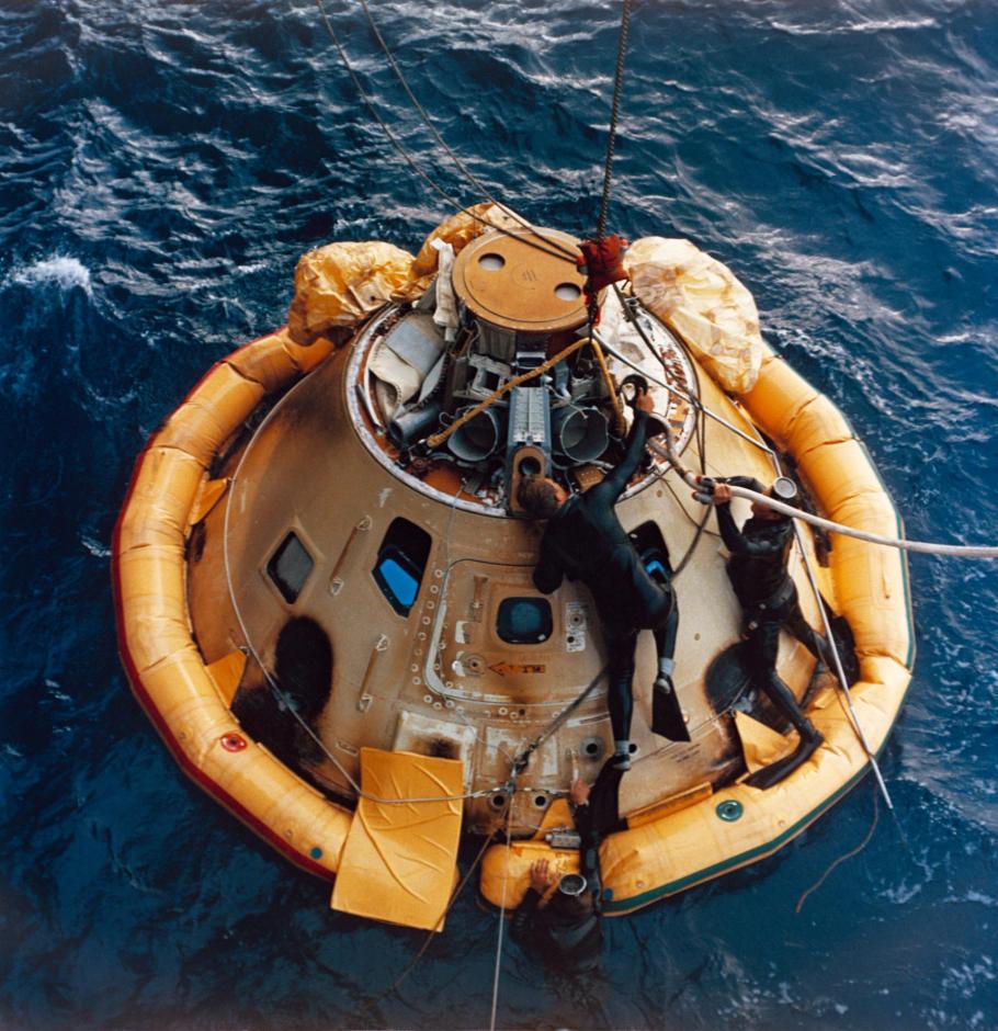 A color photograph of navy divers recovering the Apollo 6 Command Module from the Pacific Ocean. The Command Module floats on the surface of the water.