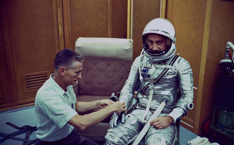 man in silver spacesuit with another man assists him with his glove