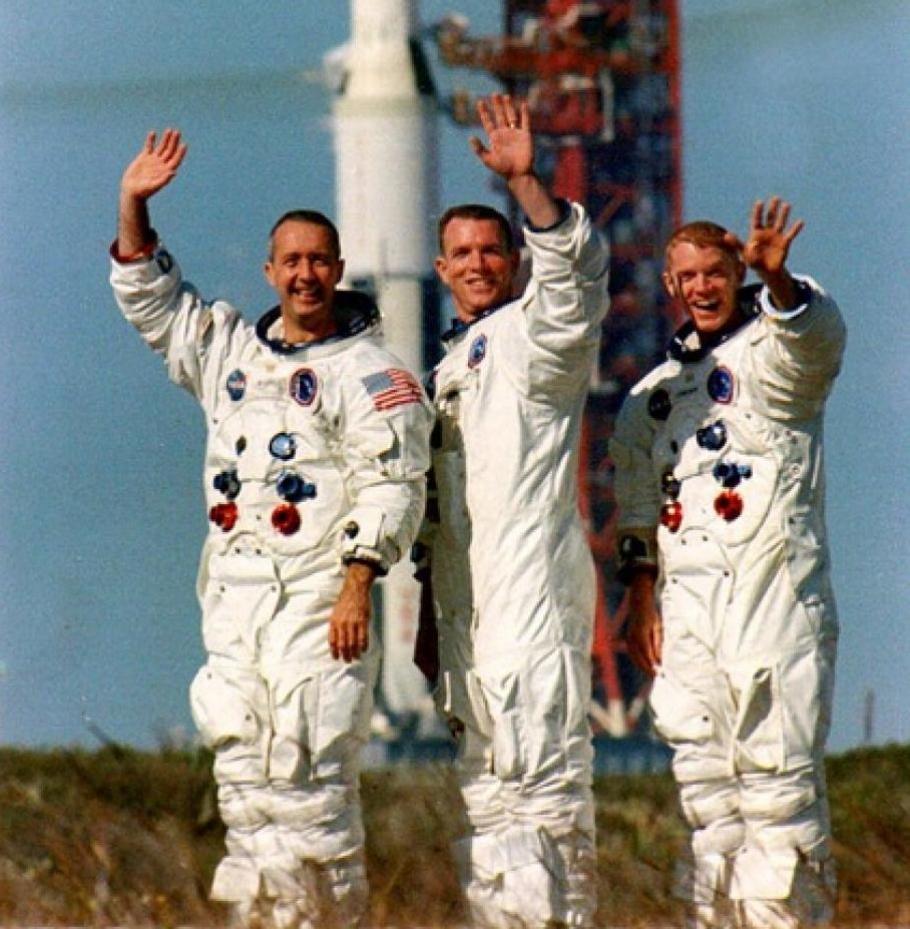 A photograph of the three men who make up the Apollo 9 crew in front of a rocket. All are waving to the camera.