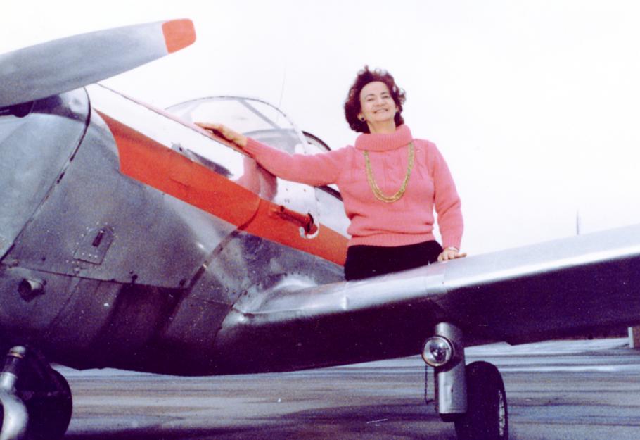 A woman in a pink sweater with amputated legs smiles while sitting on the wing of an airplane. 