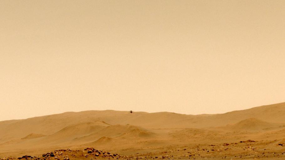 A view of the orange surface of Mars with hills in the background and a barely visible device flying above in the air.