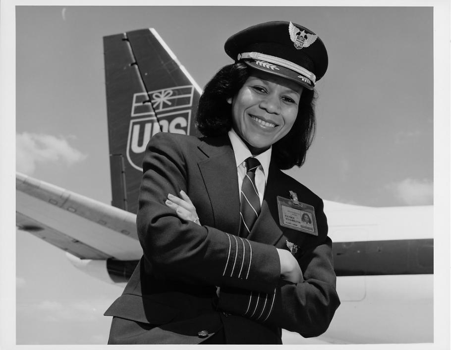 Black & white photograph. African-American woman with hair to shoulders turned to her right with left hand crossed under right.  She is wearing a dark uniform with three pale thin stripes above each hand and an ID badge on her left chest.  She has a white collared shirt with a dark tie with thin pale stripes under the hack. She wears a stiff pilots cap with two laurel leaves on the brim and a star surrounded by wings on the crest.  In the background left is an airplane tail with the UPS logo of wrapped pack