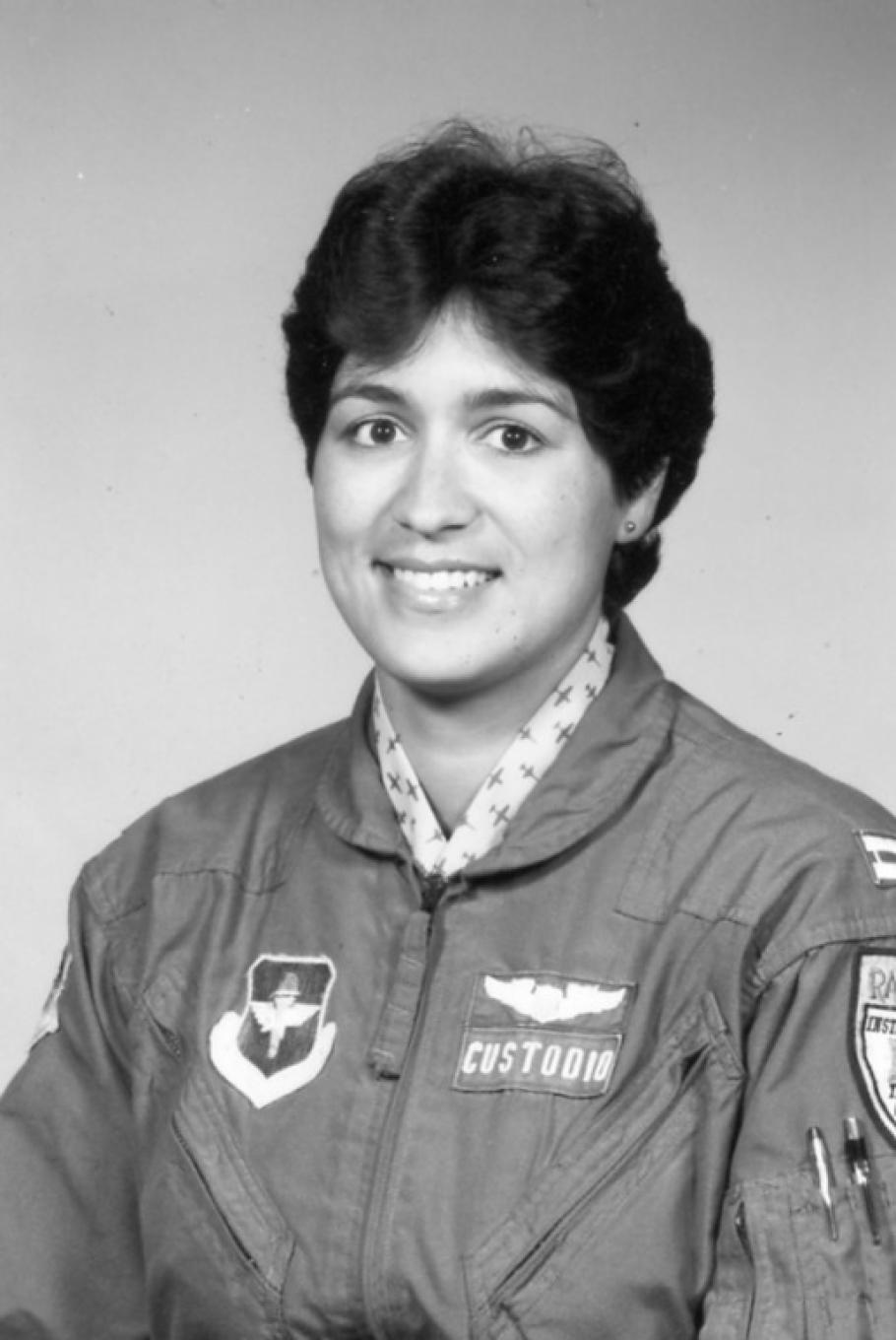 Black and white portrait of woman with short wavy hair, parted in the middle.  Stud earring in left ear (right ear is not visible).  She is wearing a dark jacket with a zipper with a cloth pull.  Patch on her right side (the left of the image) features a icon with wings.  Two patches on her left side (right of photo). Top is wings. Bottom is a nameplate that says "Custodio."  There are two pens in her left sleeve. Another patch is cut off by the edge. White collar with a dark airplane print under the jacket