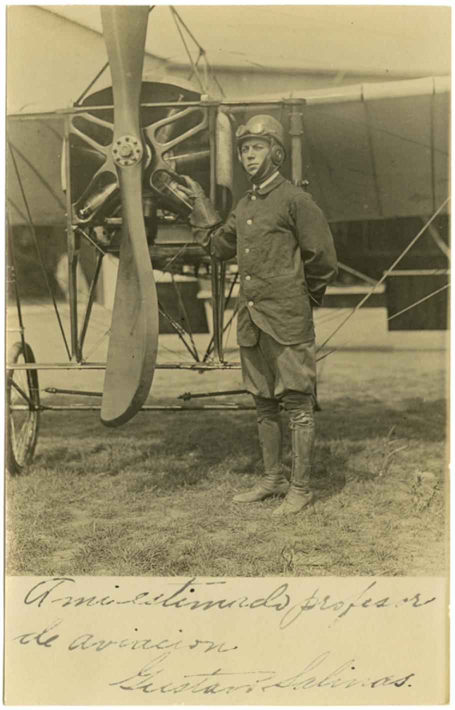 Man stands to the right of a two-bladed propeller with the blades running from the sky to the ground. He is wearing a leather cap with goggles pushed up over the cap and a button down jacket.