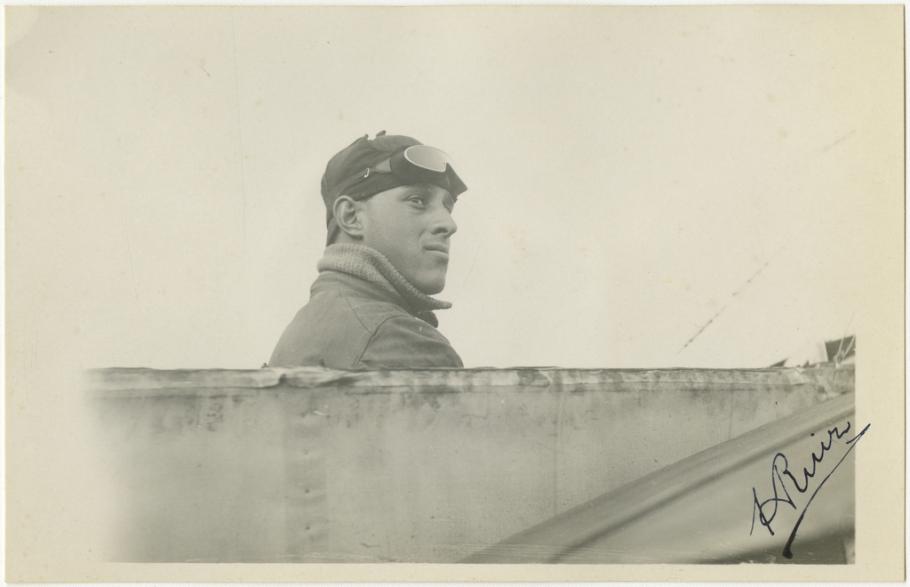 Black and white photo: Right side profile of a man (sitting in a cockpit but you can only see a line across the bottom of the image). Man is wearing a high collared jacket, leather flying cap and goggles pushed up over the cap.